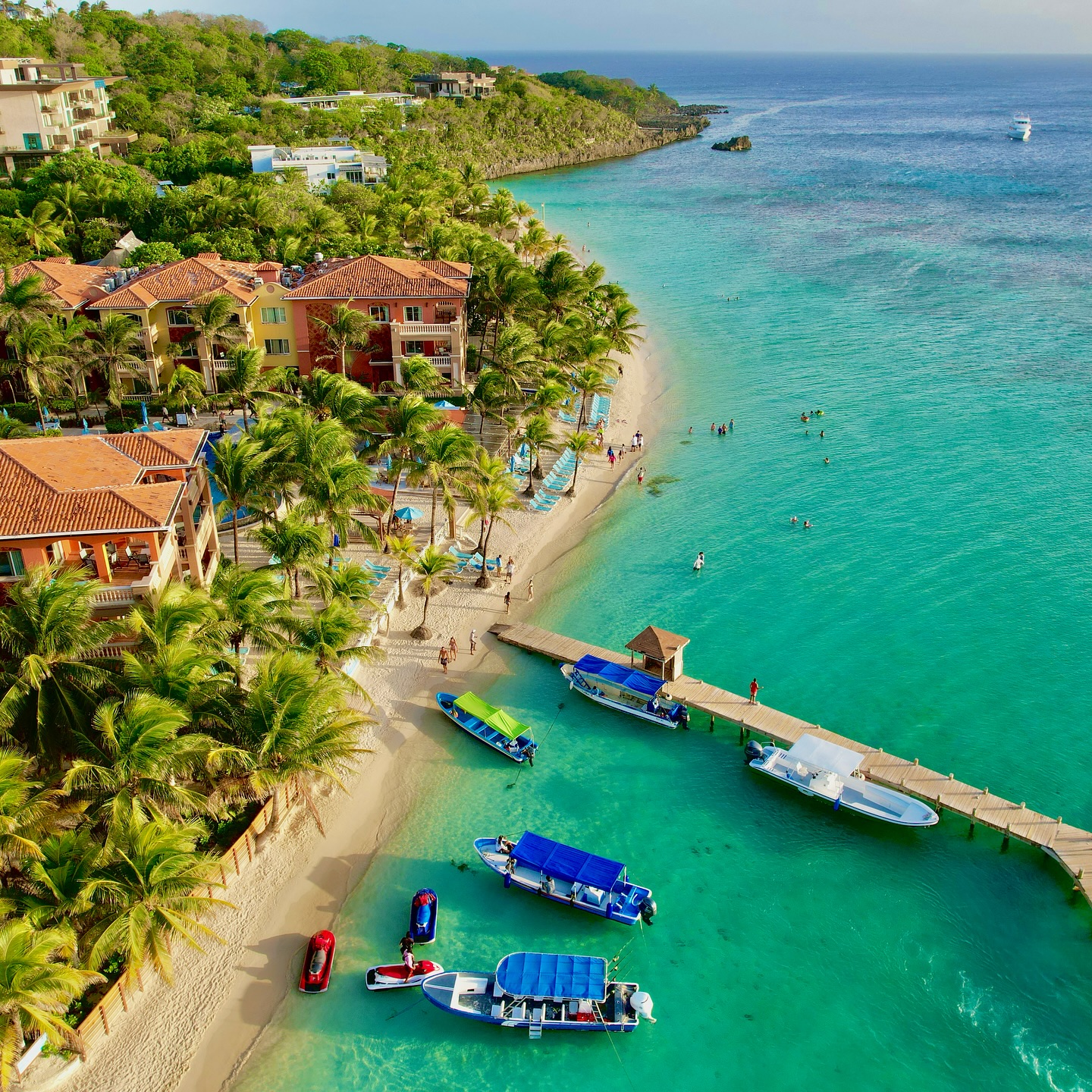 Aerial view of a vibrant Roatán beach with turquoise water, palm trees, boats and a wooden dock